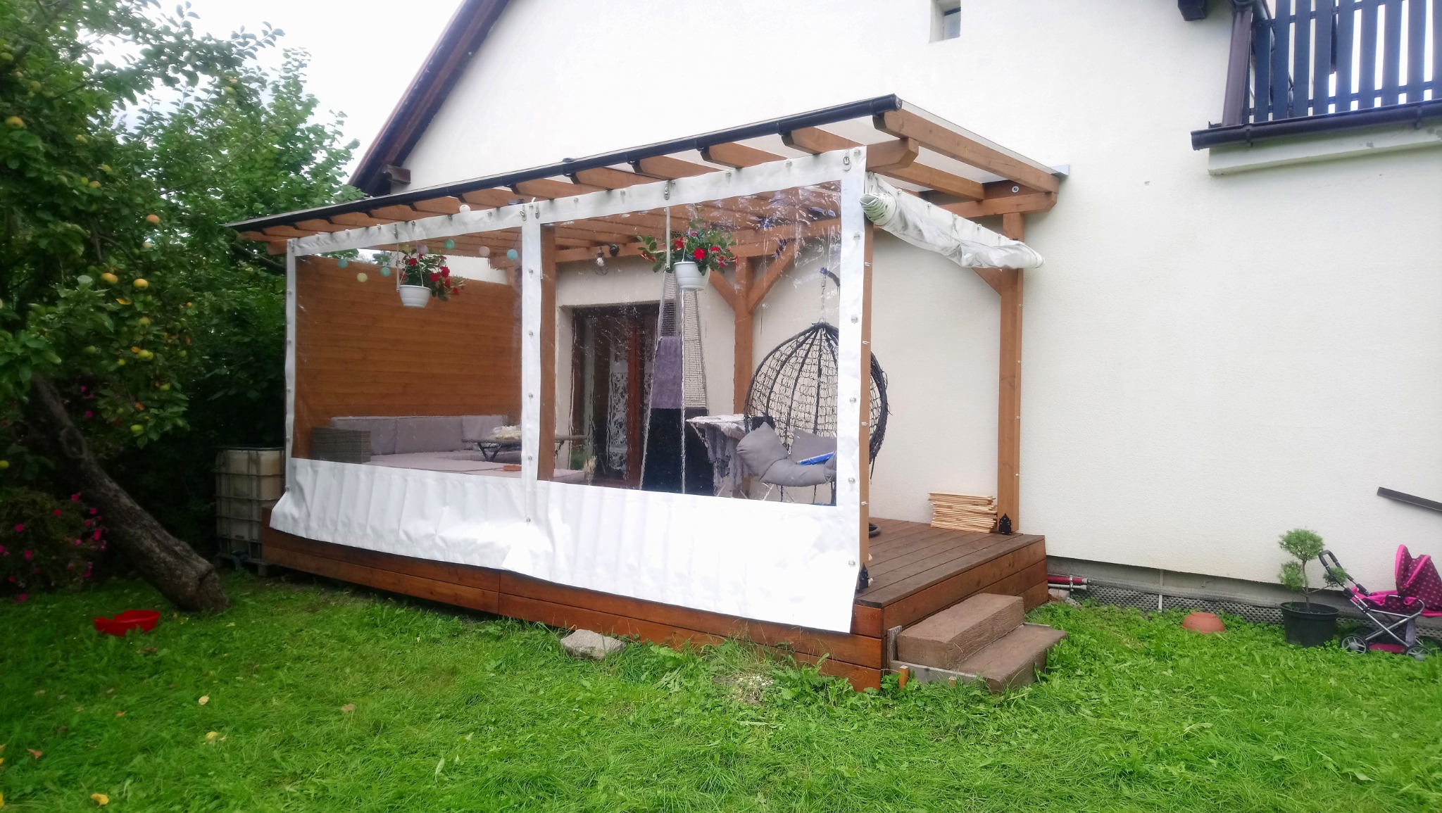 Zadaszony drewniany taras z przezroczystymi osłonami bocznymi, wyposażony w wiszący fotel i sofę, widok z zewnątrz na tle zielonego trawnika i drzew