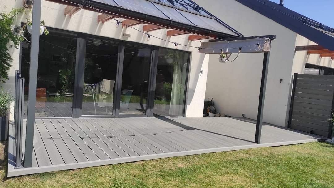 Szary taras kompozytowy przylegający do nowoczesnego domu z dużymi przesuwnymi drzwiami, oświetlony girlandą żarówek, z widocznym fragmentem szarego ogrodzenia.