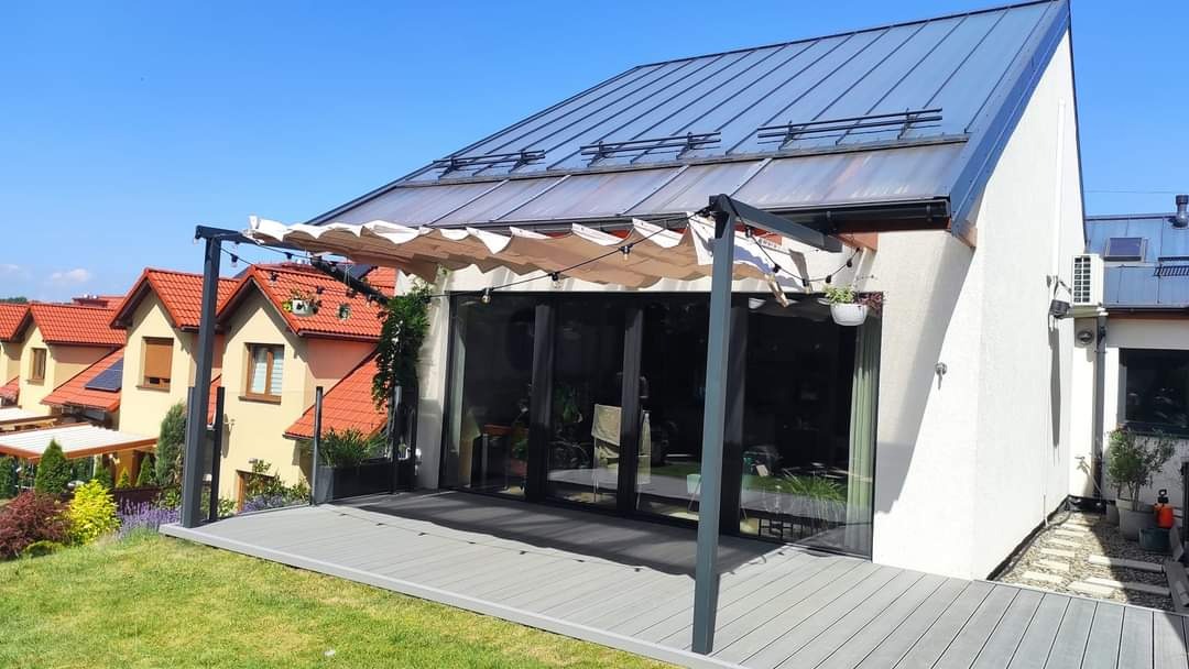 Nowoczesny taras kompozytowy w odcieniach szarości z pergolą metalową i rozsuwanym zadaszeniem tekstylnym, przylegający do budynku z dużymi przesuwnymi drzwiami szklanymi, w tle zabudowa szeregowa...