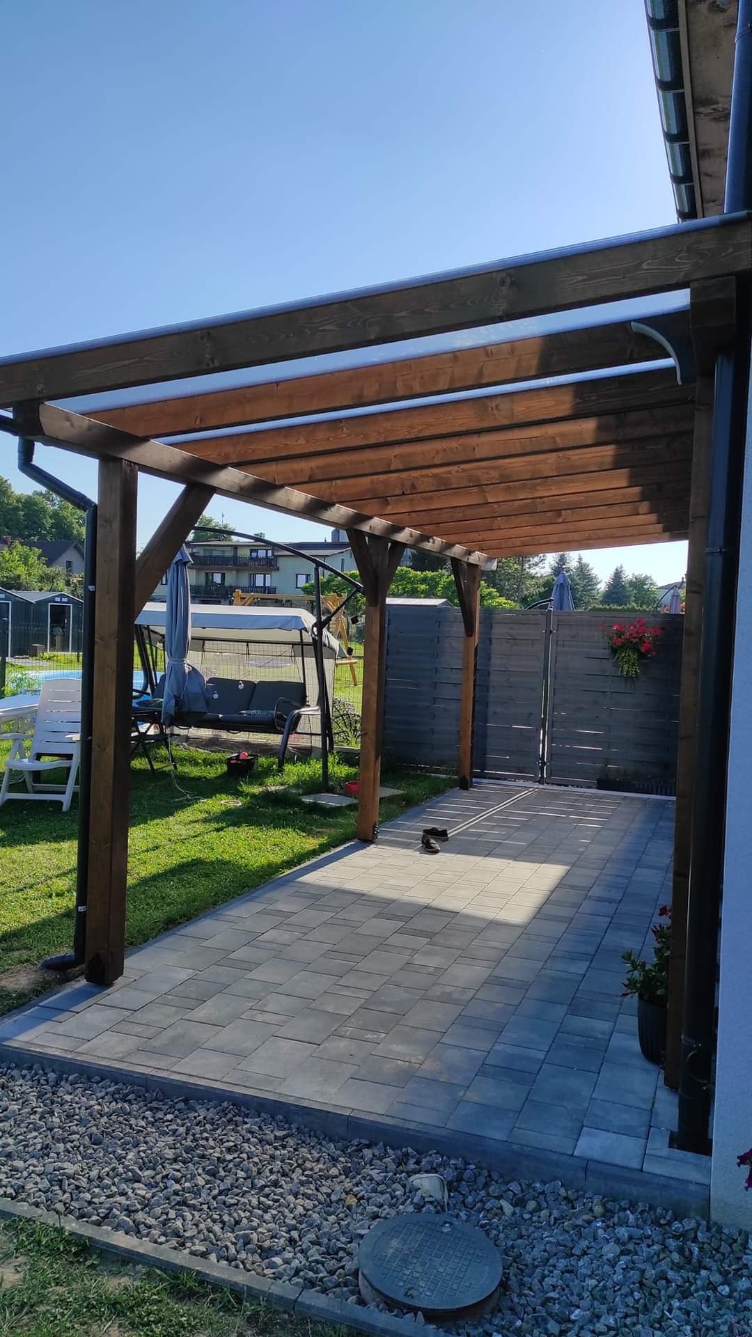 Drewniana konstrukcja z zadaszeniem z przezroczystego tworzywa, tworząca zacienione miejsce do siedzenia na wyłożonym kostką brukową tarasie, z widokiem na ogród z huśtawką i kwiatami.