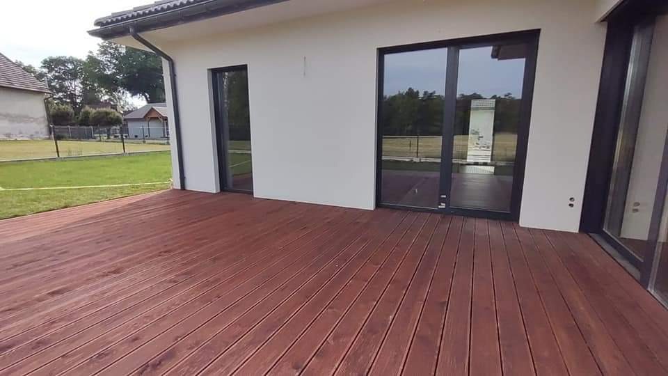 Ciemnobrązowy taras z desek przy białym domu z dużymi, przesuwnymi oknami i zielonym trawnikiem w tle.