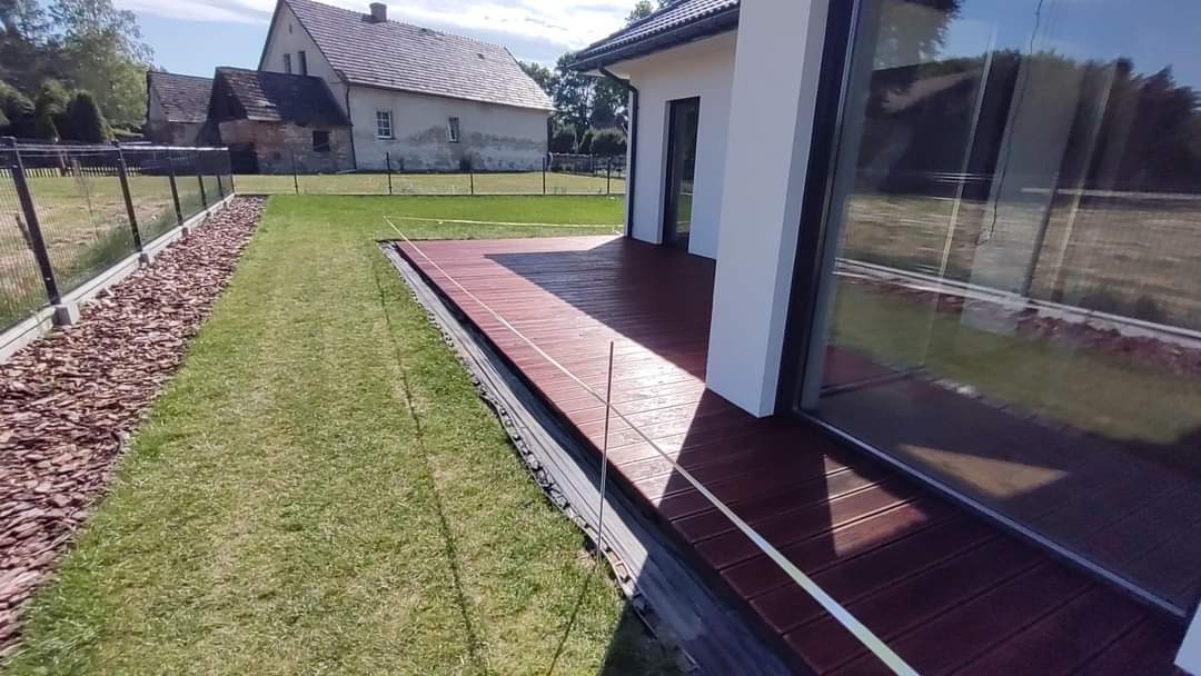 Nowoczesny taras z desek kompozytowych w kolorze ciemnego drewna, przylegający do budynku z dużymi oknami, widok na trawnik i dom w tle, widoczne linki wyznaczające poziom.