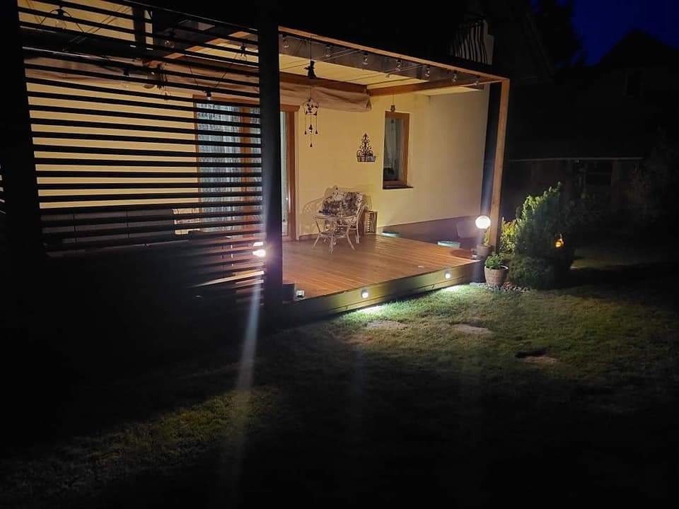 Oświetlony taras z drewnianą podłogą i pergolą z żaluzjami w ogrodzie nocą, widoczne lampy punktowe w podłodze tarasu i dekoracje.