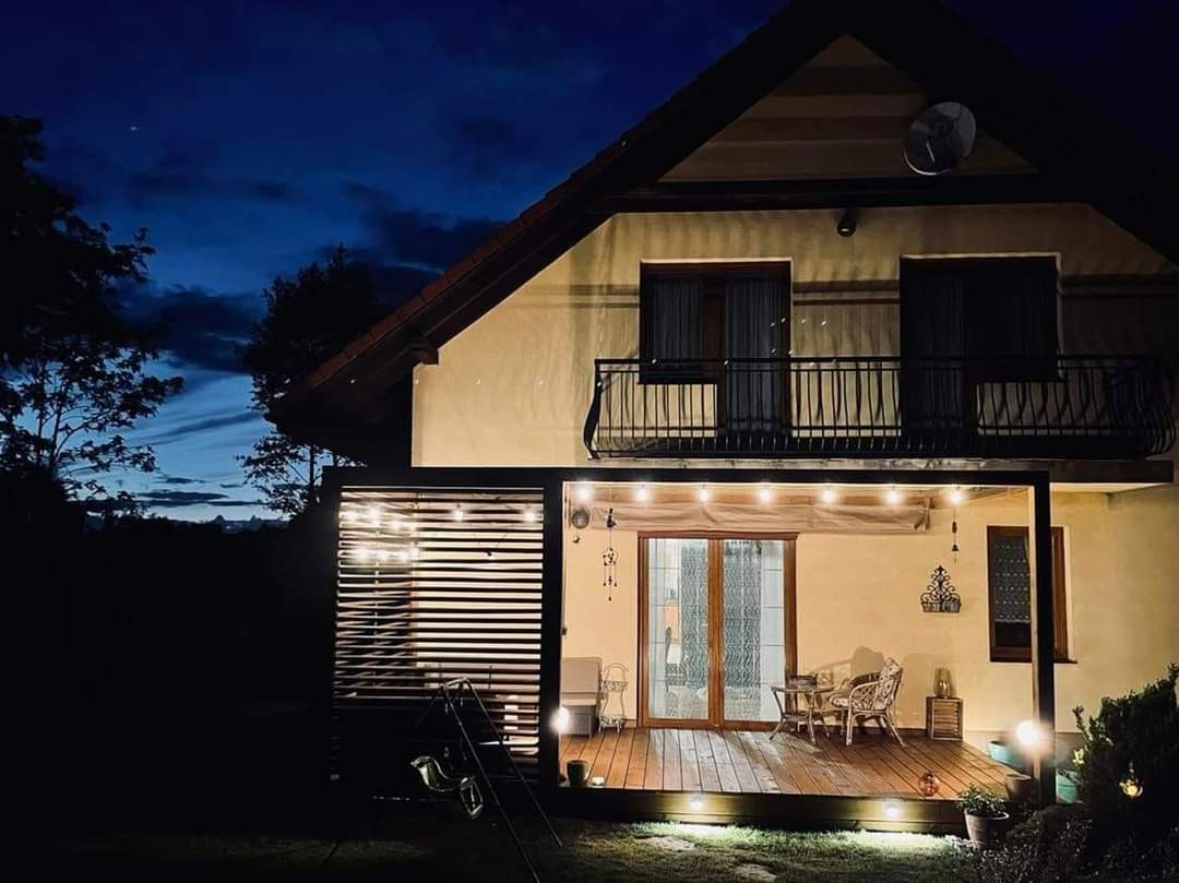 Oświetlony taras drewniany przy domu jednorodzinnym, widok wieczorny z dekoracyjnymi lampkami i meblami ogrodowymi.