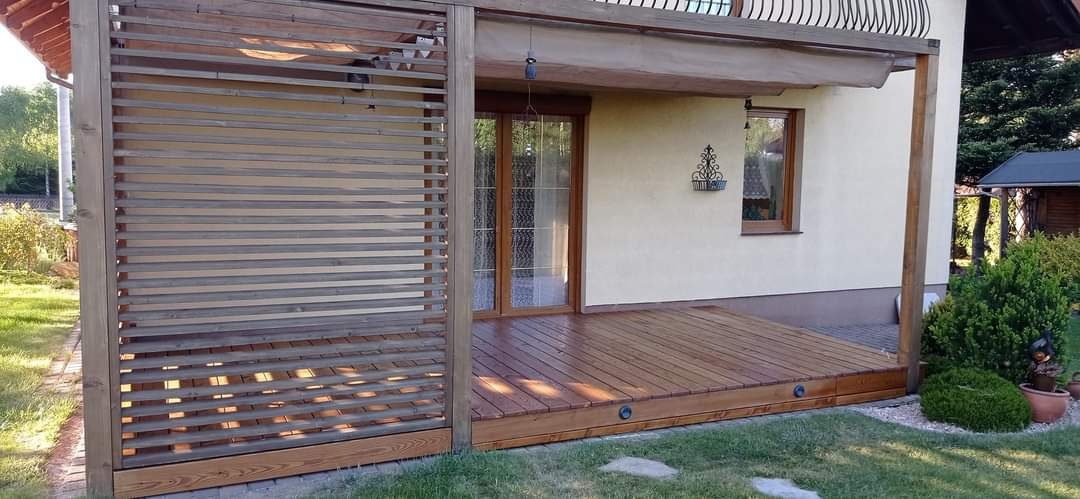 Zadaszony taras z drewnianą podłogą i ażurową ścianą, zintegrowany z budynkiem mieszkalnym, oświetlenie punktowe w podłodze, widok na ogród.