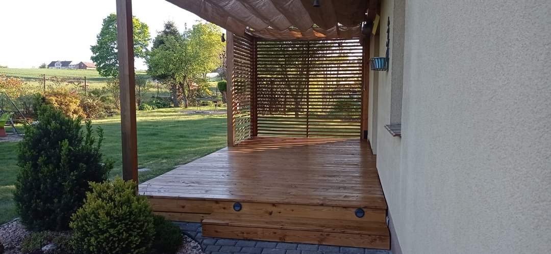 Zadaszony taras drewniany z ażurową ścianą, oświetleniem punktowym w podstopnicach i beżową tkaniną zacieniającą nad głową, widok na zielony ogród w tle.