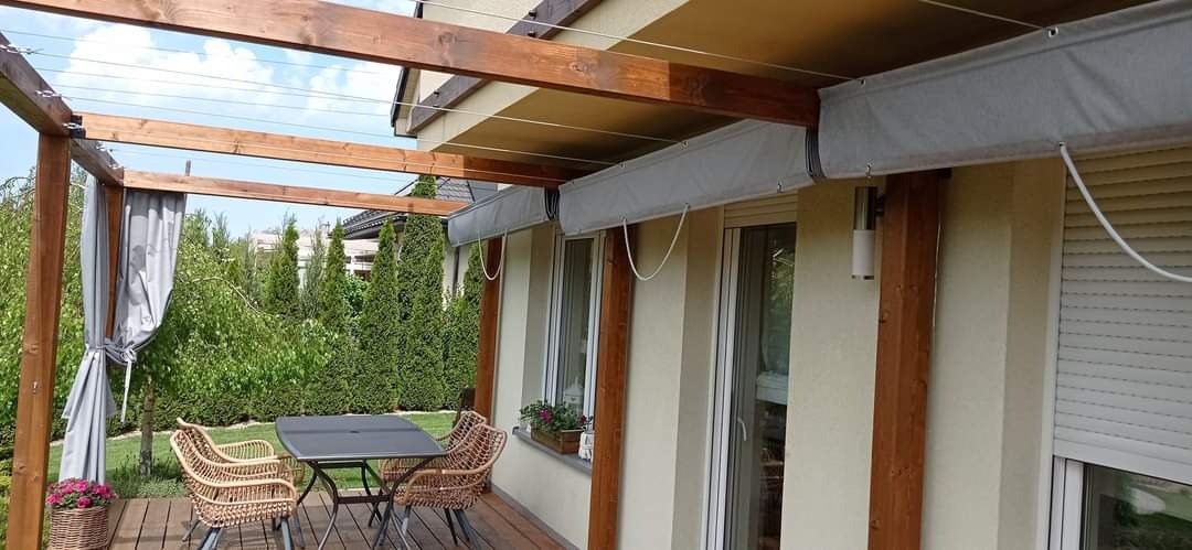 Drewniana pergola z zasłonami i roletami przeciwsłonecznymi, rattanowe krzesła i stolik na drewnianym tarasie, widok na zielony ogród.