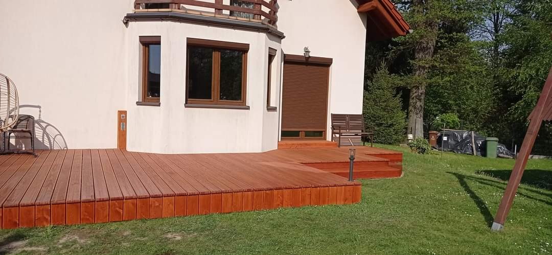 Nowoczesny, brązowy taras z drewna otaczający dom z beżową elewacją i brązowymi oknami, widok na trawnik i drzewa w tle.