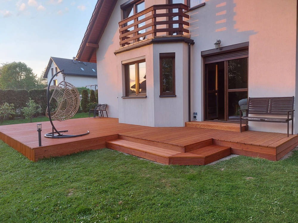 Nowoczesny taras z drewna egzotycznego na trawniku, z wiszącym fotelem i schodkami prowadzącymi do domu, oświetlony ciepłym światłem zachodzącego słońca.