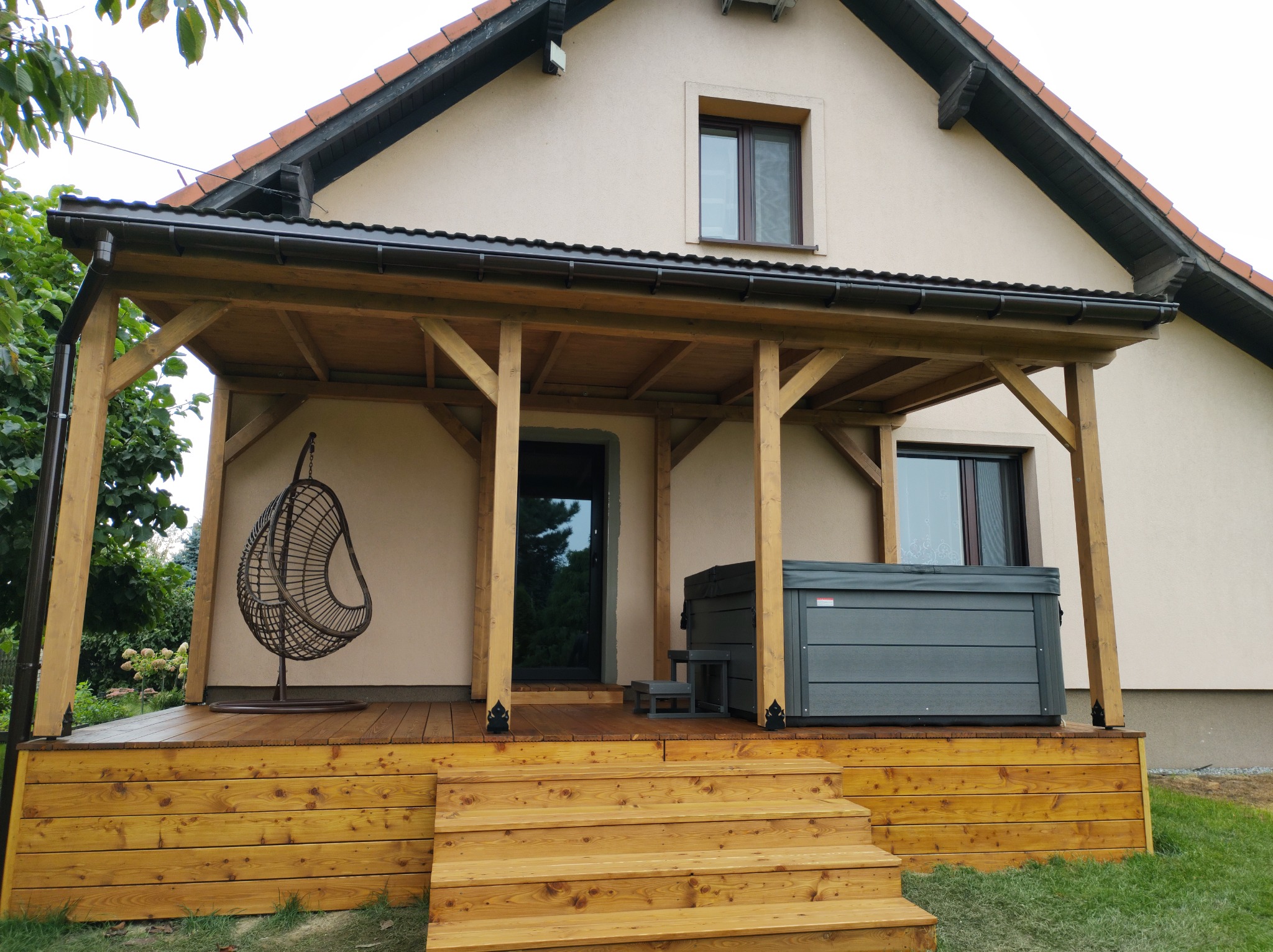 Zadaszony taras z drewna z jacuzzi i wiszącym fotelem na tle domu jednorodzinnego.