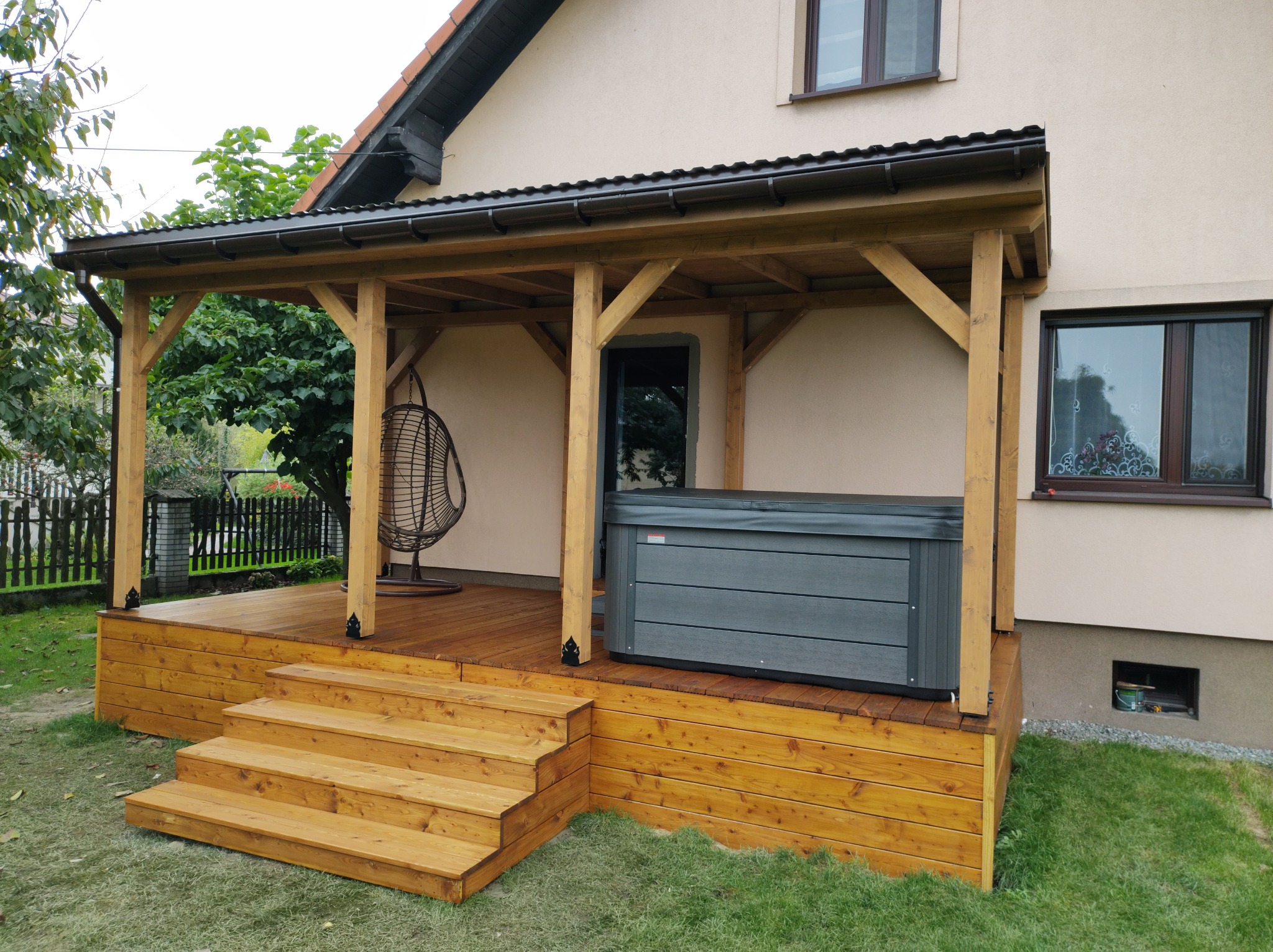 Zadaszony taras z drewna z zewnętrznym jacuzzi, widoczne schody wejściowe, drewniane słupy podtrzymujące zadaszenie i wiszące krzesło kokon w tle.