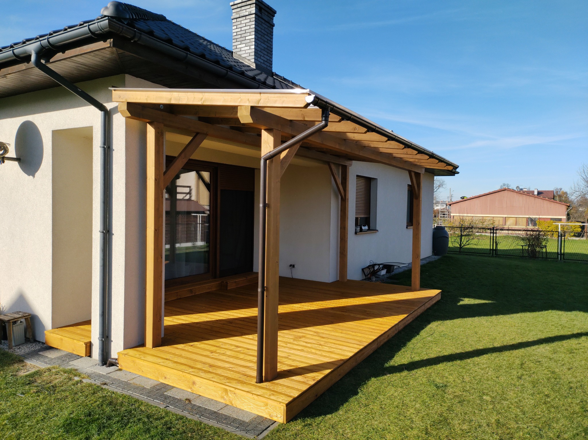 Drewniany taras przylegający do domu, z zadaszeniem wspartym na drewnianych słupach, widok na zadbany trawnik i błękitne niebo.