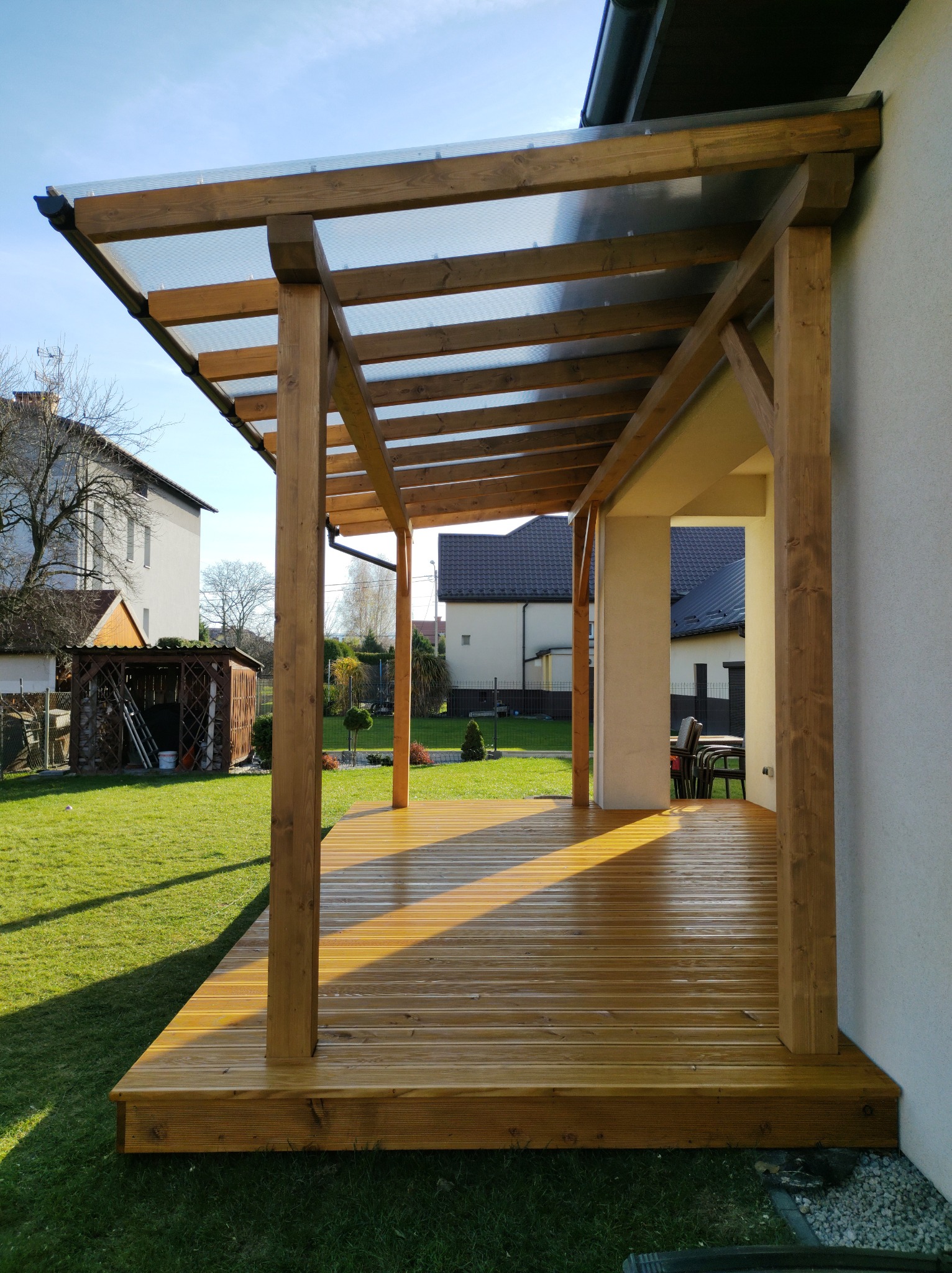 Zadaszony taras z drewna z przezroczystym zadaszeniem, widok na ogród z domkami i zielony trawnik w słoneczny dzień.