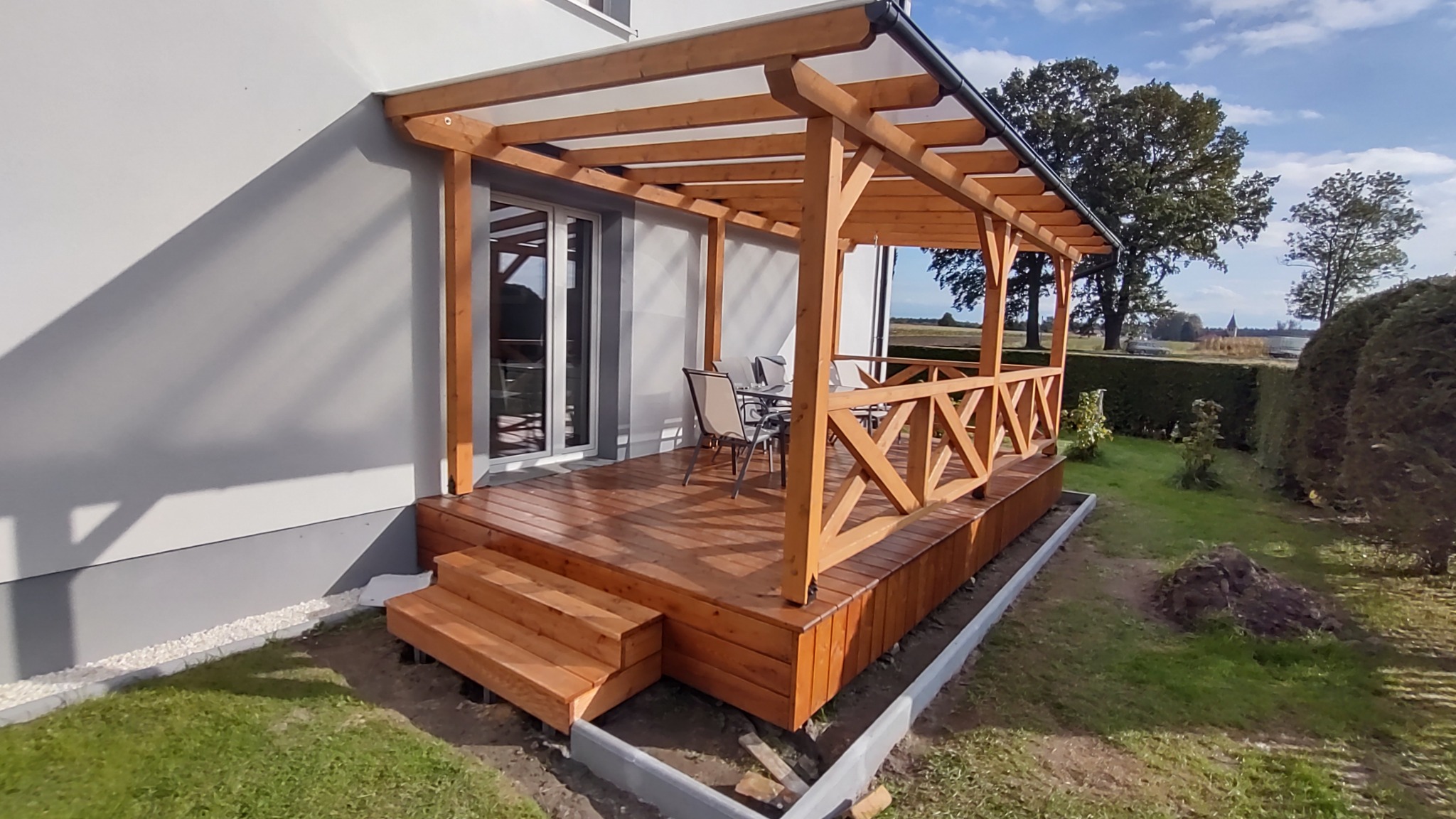 Drewniany taras z zadaszeniem z poliwęglanu, balustradą z krzyżowych belek i schodami, przylegający do szarego domu z przesuwnymi drzwiami tarasowymi, na trawniku z widokiem na drzewa w oddali.