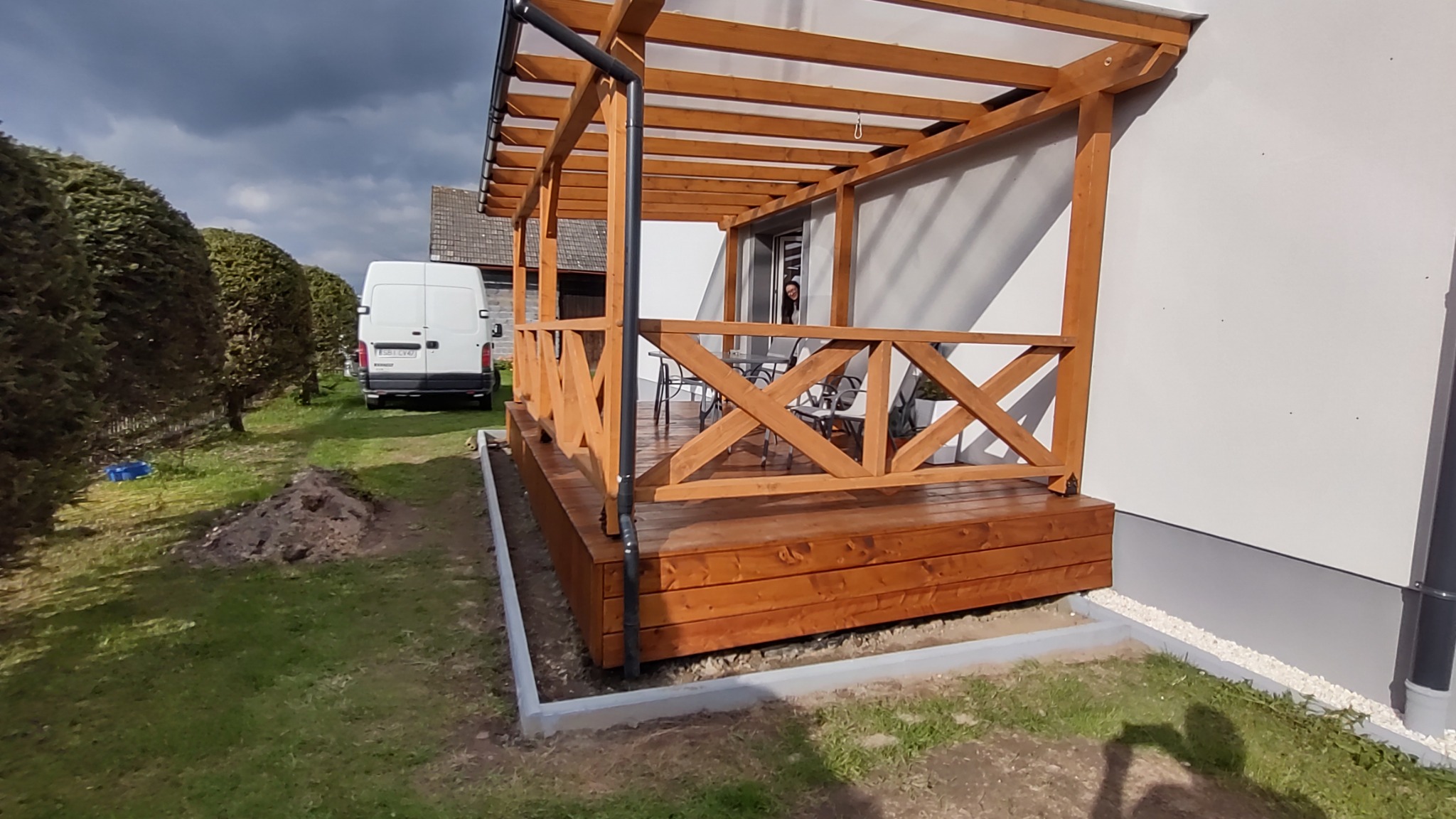 Zadaszony taras z drewnianą konstrukcją, balustradą z krzyżowych elementów i drewnianą podłogą, widok z zewnątrz na tle domu i zieleni.