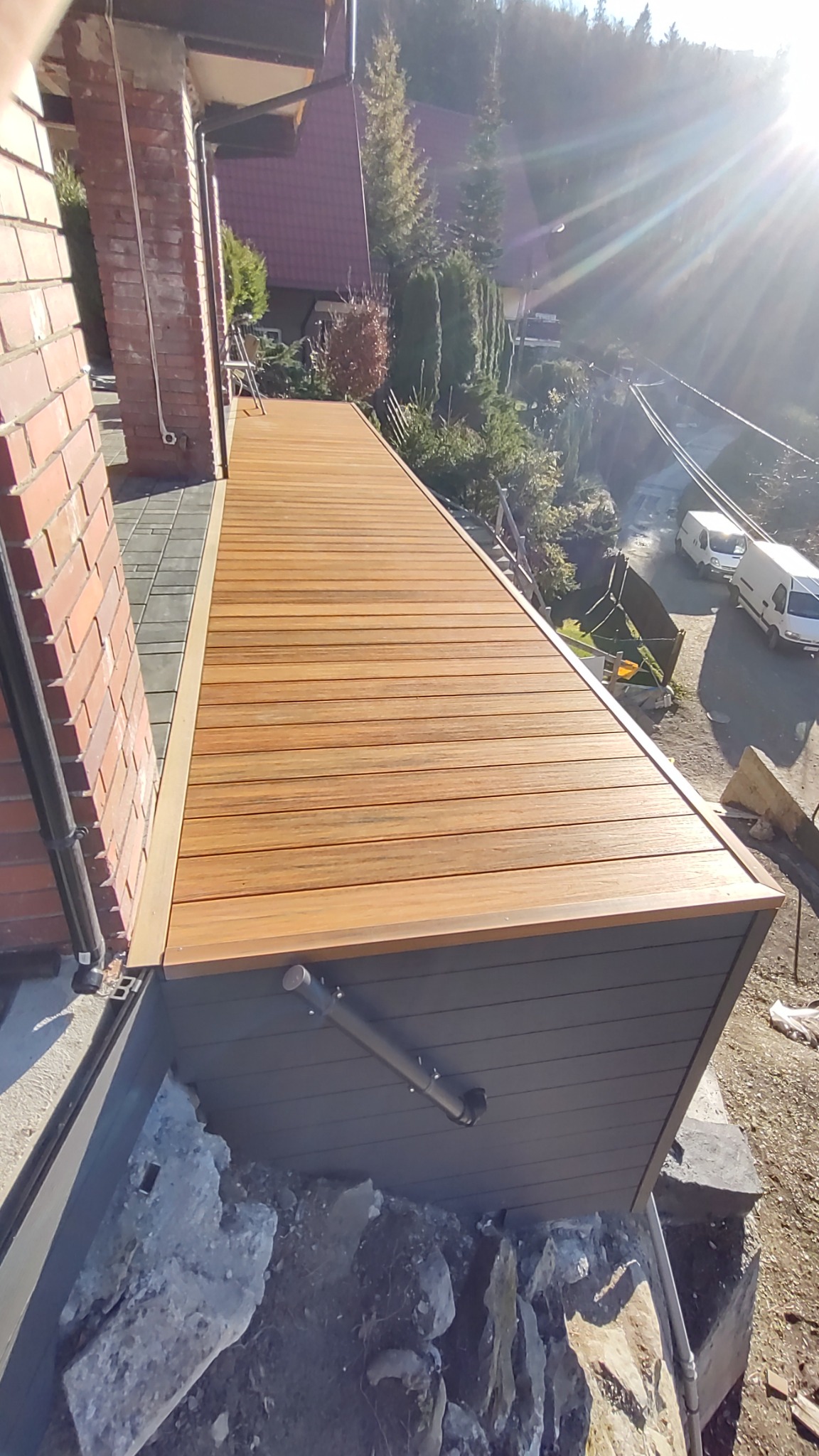 Nowoczesny taras z desek kompozytowych w odcieniach brązu, przylegający do ceglanego budynku, widok z góry na otoczenie z domami i drzewami w słoneczny dzień.