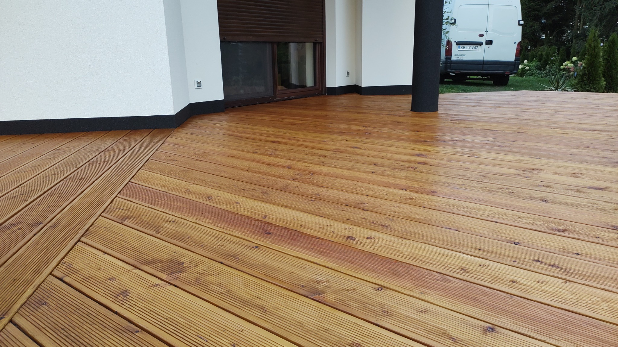 Świeżo zaimpregnowany taras z desek drewnianych, widok z niskiej perspektywy, fragment ściany budynku z oknem i zaparkowany biały bus w tle.