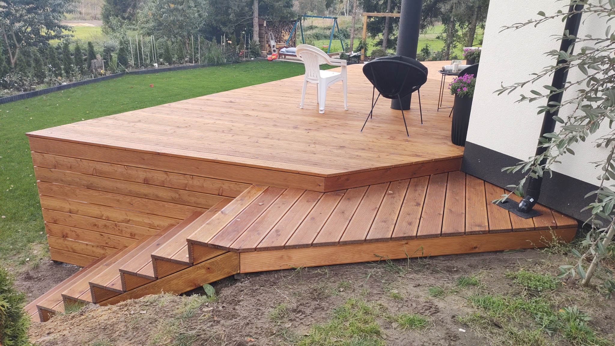 Nowoczesny taras z drewna z niskimi schodami, otoczony zielonym trawnikiem i drzewami, z widocznym kominem i meblami ogrodowymi.