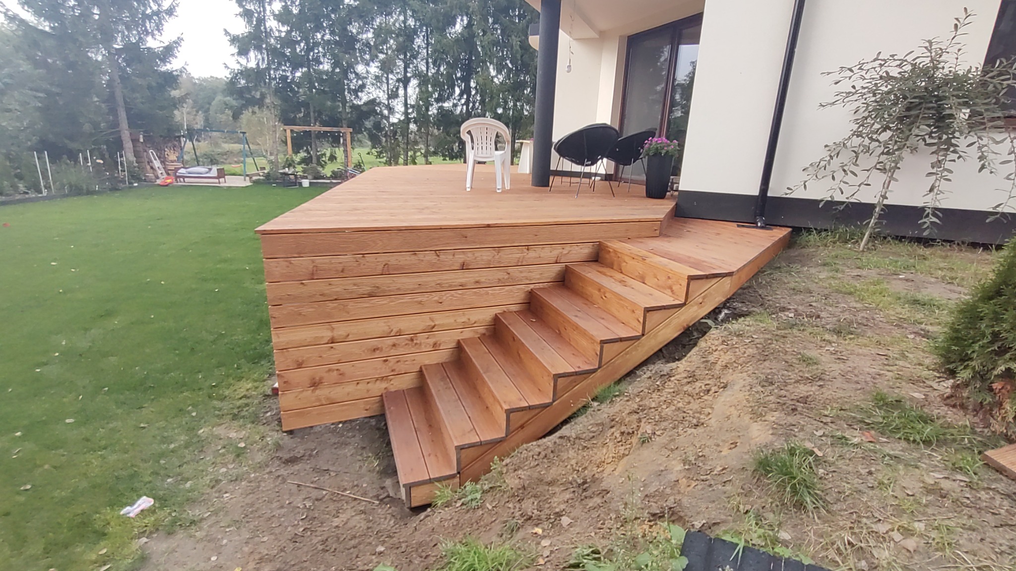 Nowoczesny taras z drewna ze schodami, z widokiem na trawnik i huśtawkę ogrodową w tle, uzupełniony białym krzesłem i czarnymi fotelami.