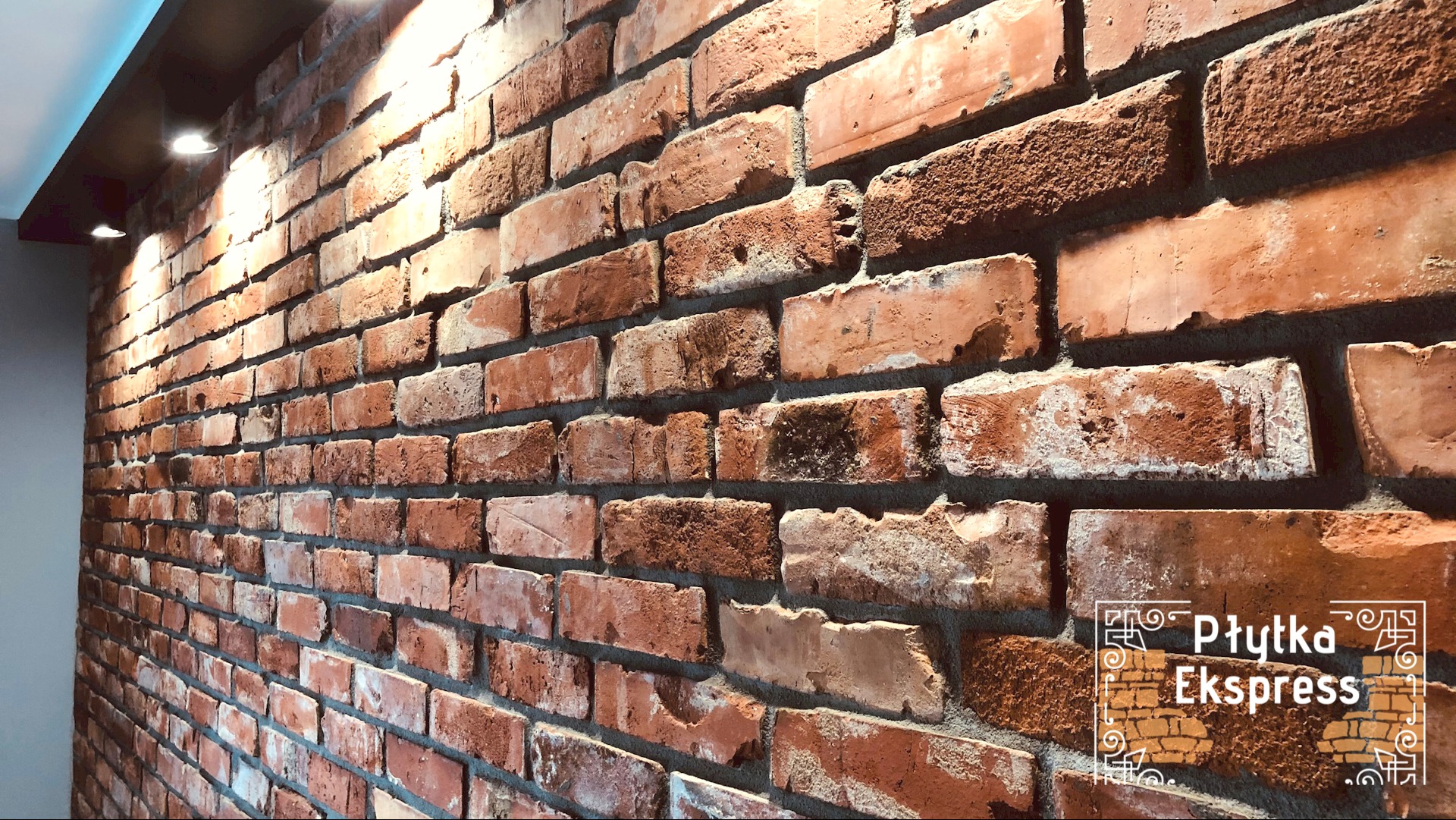 Ściana z czerwonej cegły z widocznym logo 'Płytka Ekspress' w prawym dolnym rogu.