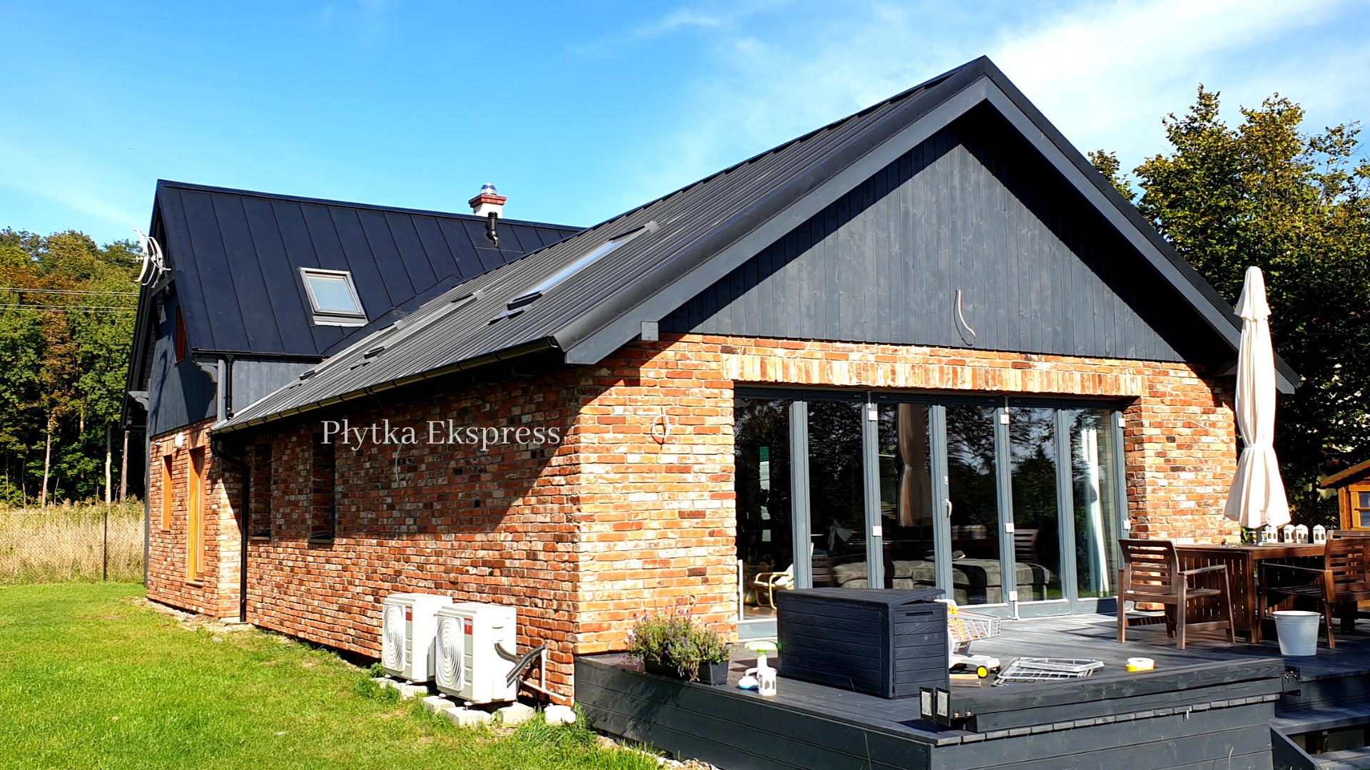 Nowoczesny dom z cegły klinkierowej z czarnym dachem i stolarką okienną, widoczne klimatyzatory na zewnątrz i taras z meblami ogrodowymi.