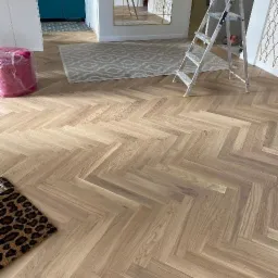 Ułożona w jodełkę jasna podłoga panelowa w Elblągu, z widoczną drabinką malarską w tle i fragmentem dywanu ze zwierzęcym wzorem.