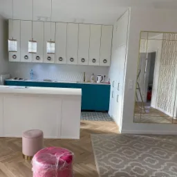 Wykończona kuchnia w stylu nowoczesnym: białe szafki z okrągłymi uchwytami, turkusowy blat, wyspa kuchenna z białym blatem, trzy wiszące lampy z przezroczystymi kloszami, parkiet w jodełkę, duże...