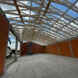 Montaż drewnianej więźby dachowej na budynku z pomarańczowej cegły, widoczni pracownicy na podnośniku i drabinie, błękitne niebo w tle