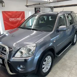 Szary samochód Honda CR-V z orurowaniem i dodatkowymi reflektorami, stojący w garażu z podłogą wyłożoną płytkami i banerami na ścianach.