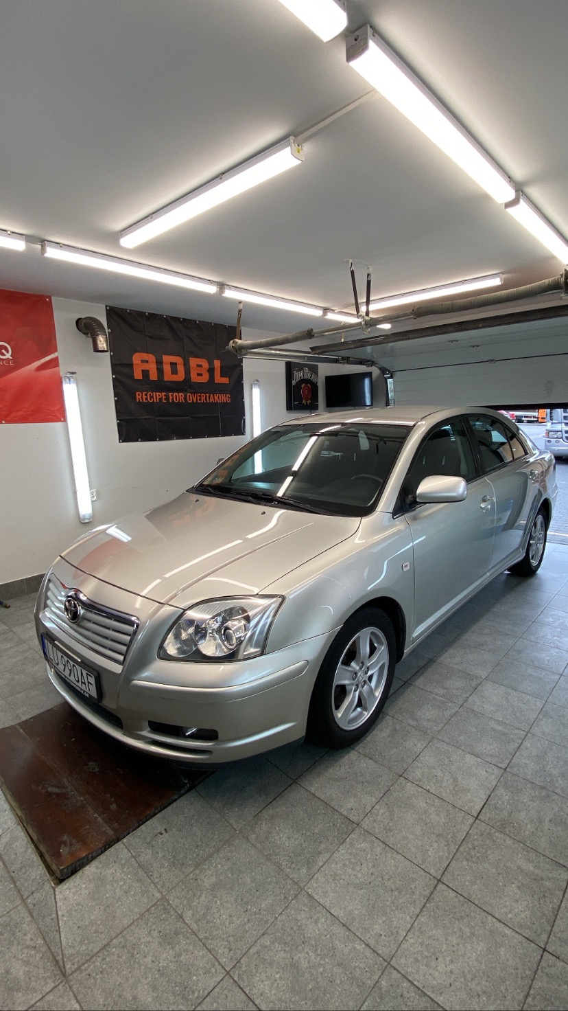 Srebrny sedan Toyota Avensis na podłodze z szarych płytek w dobrze oświetlonym garażu z banerem ADBL i otwartą bramą garażową w tle.