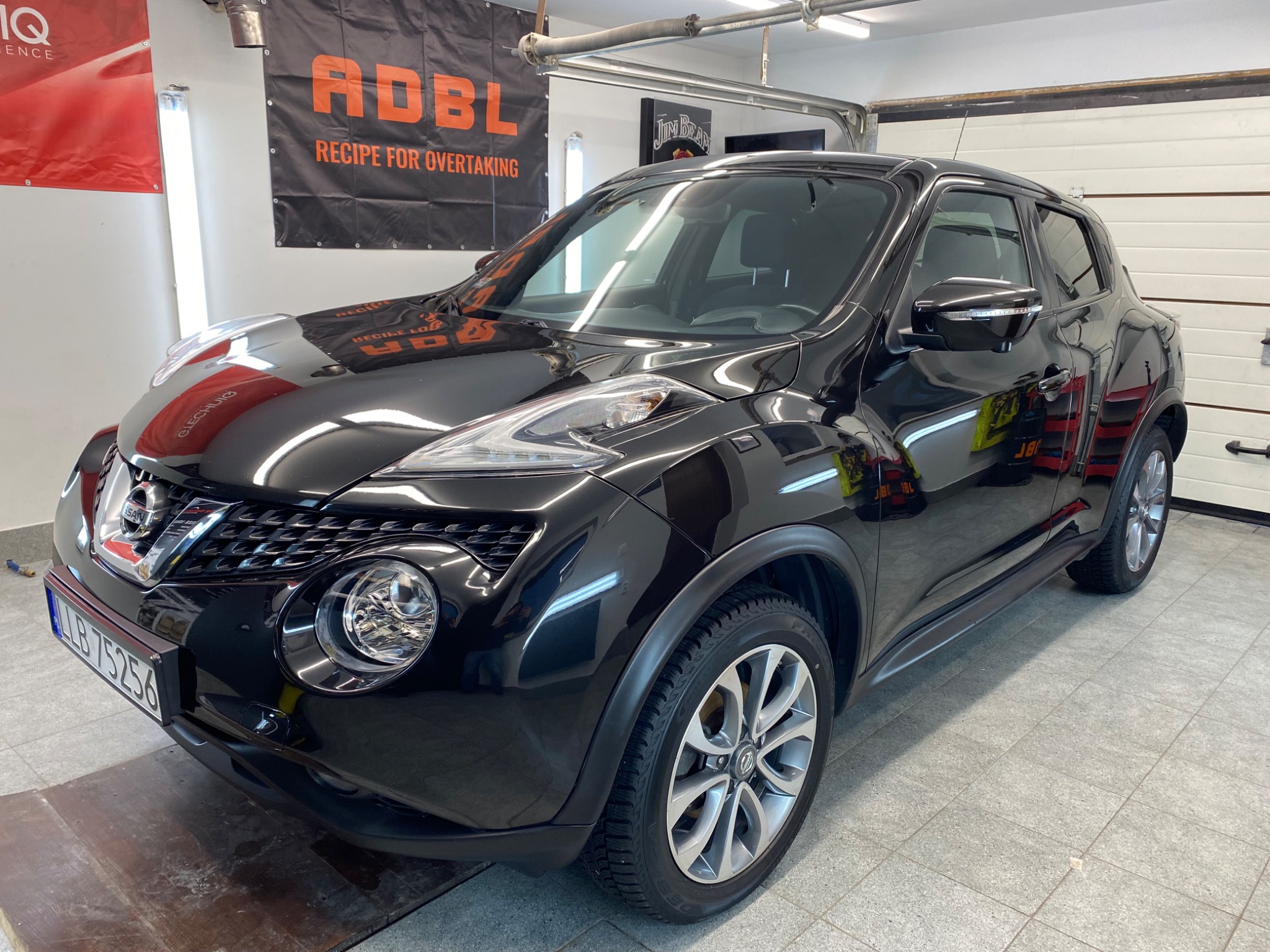 Błyszczący, czarny Nissan Juke po umyciu w myjni, widoczny z przodu pod kątem, z banerem reklamowym ADBL w tle.
