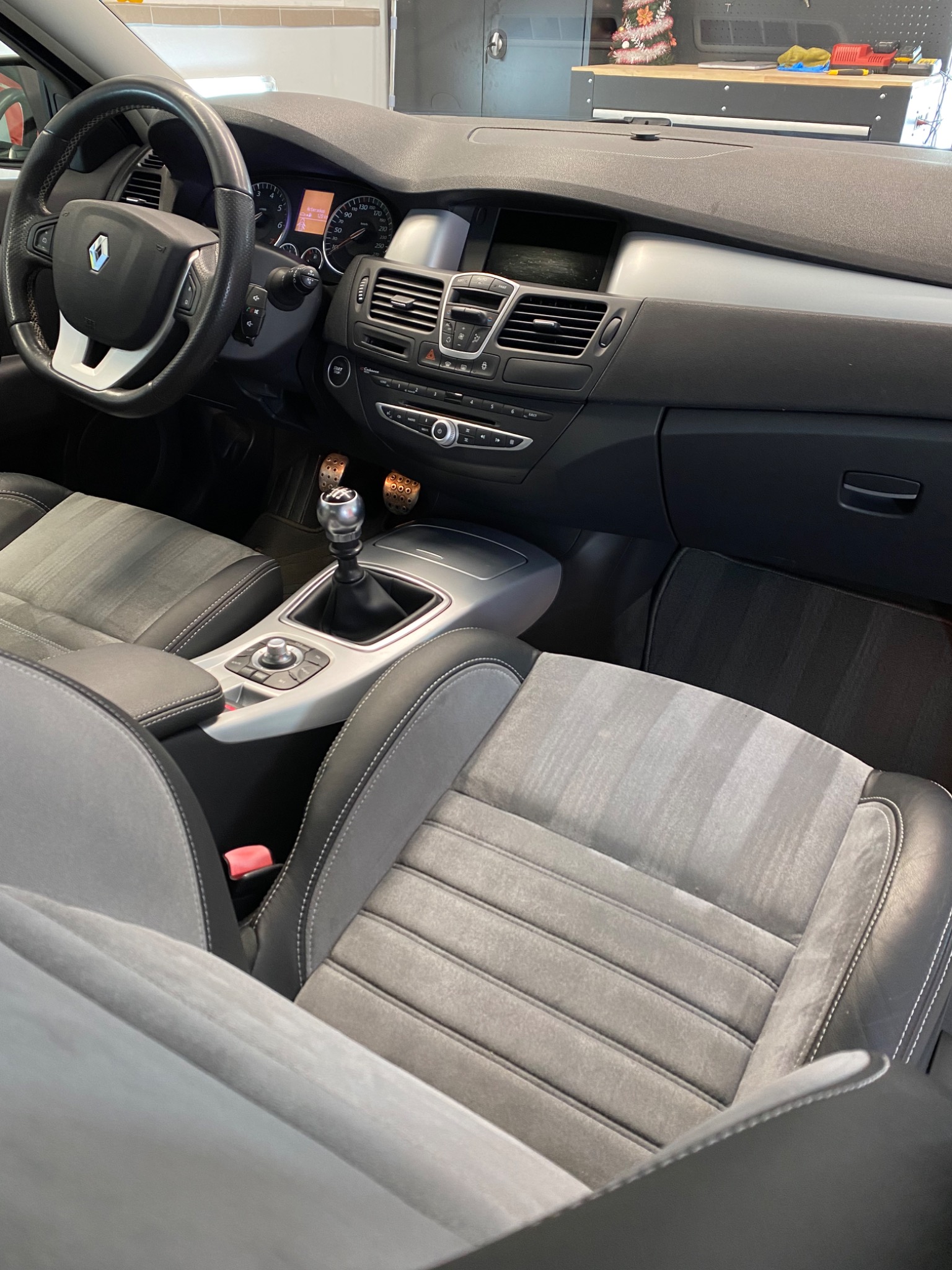 Wnętrze samochodu Renault Laguny z szarą tapicerką, widok na deskę rozdzielczą, kierownicę i fotele z wyraźnymi przeszyciami, w tle warsztat z narzędziami.