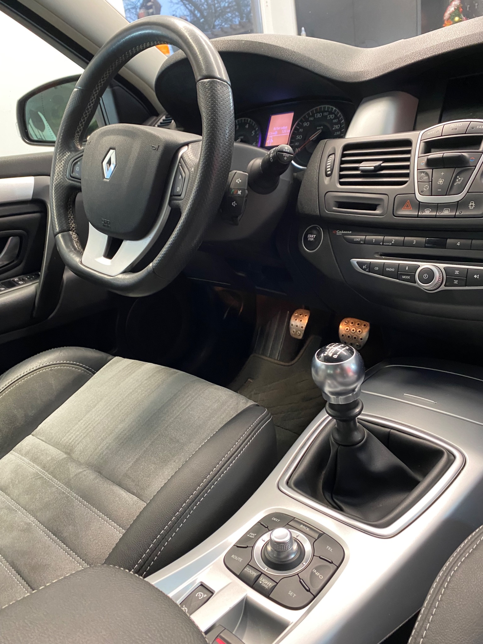 Wnętrze samochodu Renault Laguna: kierownica z logo, deska rozdzielcza z wyświetlaczem, srebrna gałka zmiany biegów, aluminiowe pedały, tapicerowane szare fotele z czarnymi obszyciami, widok...