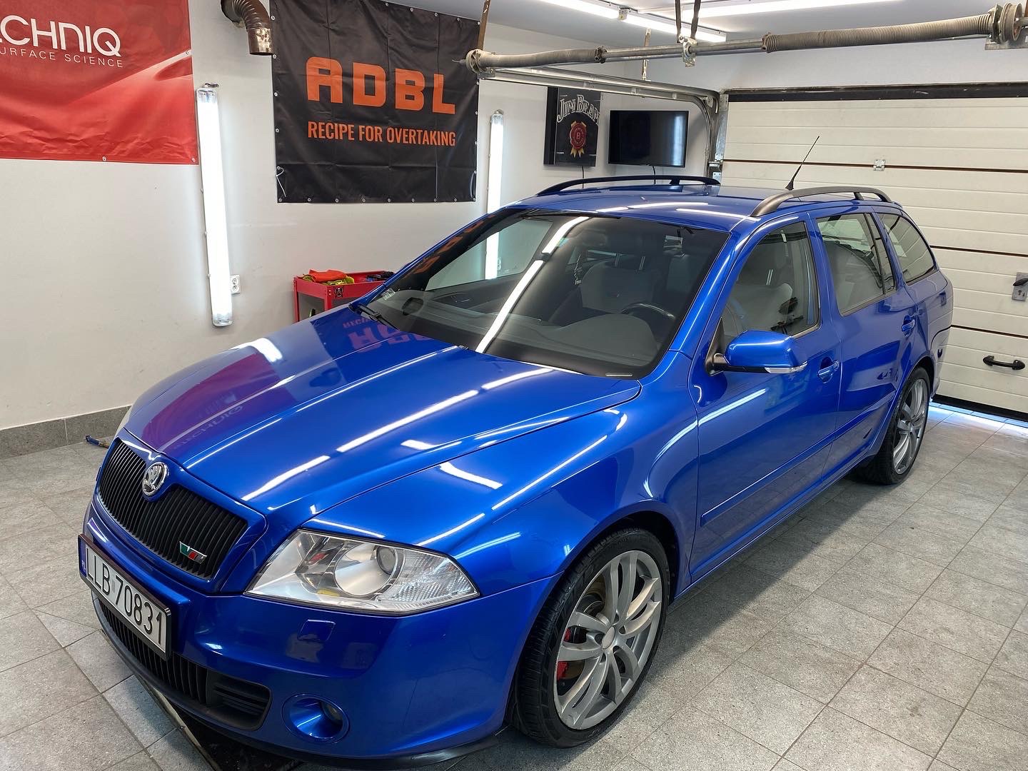 Błyszcząca, niebieska Skoda Octavia kombi po detailingu, widoczna w garażu z podniesioną bramą, z flagami RDBL i Techniq w tle.