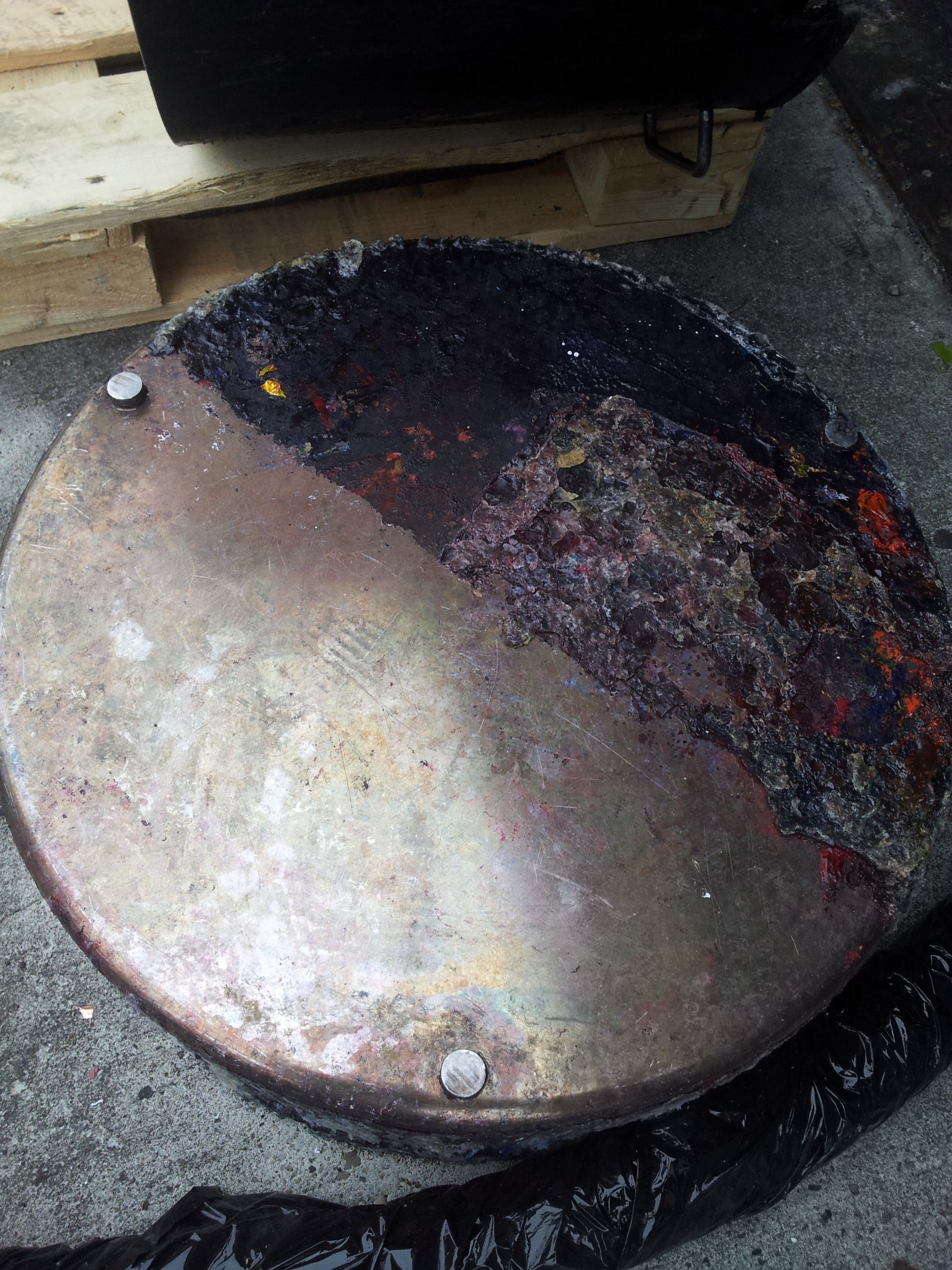 Okrągły metalowy element z wyraźnymi śladami korozji i pozostałościami po procesie, prawdopodobnie farby, z widocznymi śrubami mocującymi, leżący na betonowej powierzchni obok drewnianej palety...