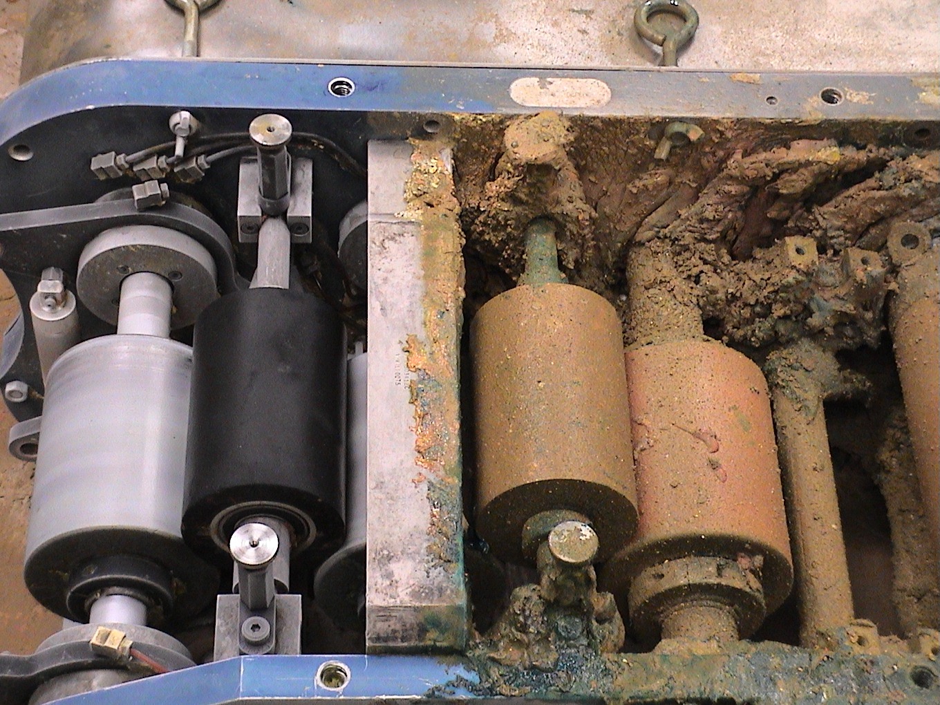 Wnętrze maszyny przemysłowej z cylindrycznymi rolkami, część z nich pokryta grubą warstwą zaschniętego brudu i zanieczyszczeń, druga część czysta, widoczne elementy hydrauliki i mocowania.