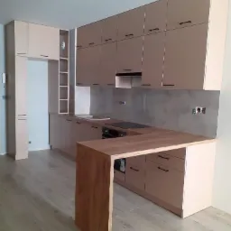 Nowoczesna kuchnia w odcieniach beżu z drewnianym blatem wyspowym, czarne uchwyty, szara ściana nad blatem roboczym, widoczne gniazdka elektryczne, minimalistyczny design, podłoga imitująca drewno.