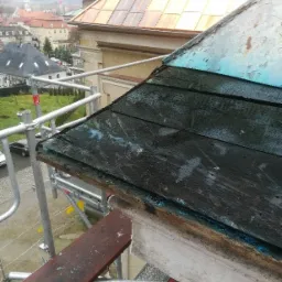 Demontaż starego, drewnianego dachu z łuszczącą się farbą podczas prac na rusztowaniu, z widokiem na miasto w tle.