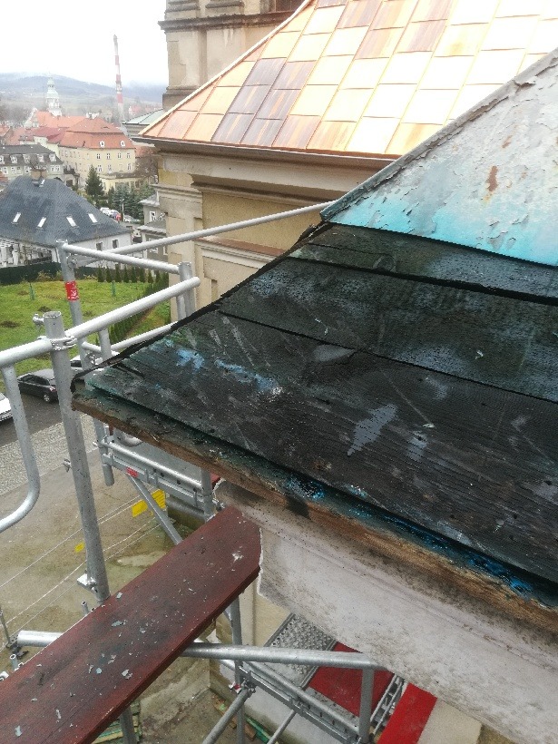 Demontaż starego, drewnianego dachu z łuszczącą się farbą podczas prac na rusztowaniu, z widokiem na miasto w tle.