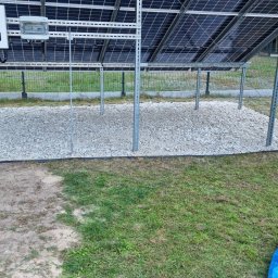 T.W. NORTMAR Power Systems - Instalacja paneli słonecznych na metalowej konstrukcji, podsypana białym kamieniem, z widocznym inwerterem i małym białym psem w ogrodzie.