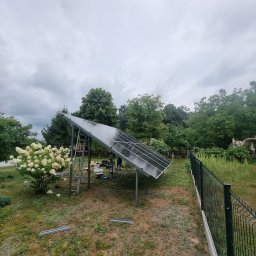 T.W. NORTMAR Power Systems - Montaż paneli słonecznych w ogrodzie: widoczne panele na metalowej konstrukcji, drabina, narzędzia, trawa i kwiaty hortensji, pochmurne niebo w tle.