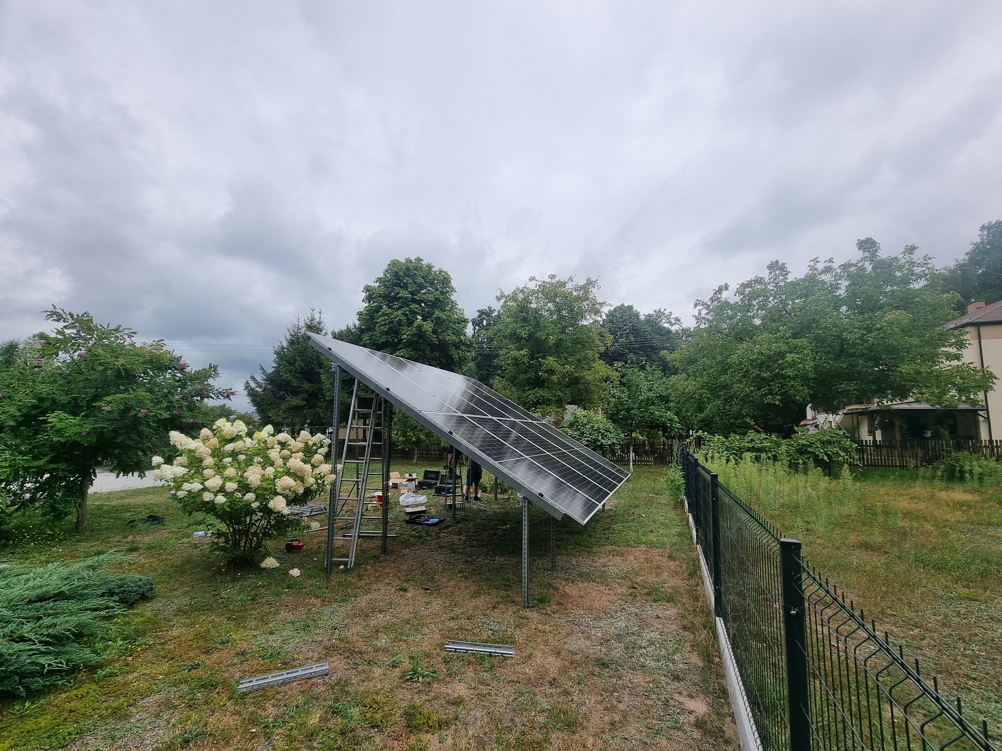 Montaż paneli słonecznych w ogrodzie: widoczne panele na metalowej konstrukcji, drabina, narzędzia, trawa i kwiaty hortensji, pochmurne niebo w tle.