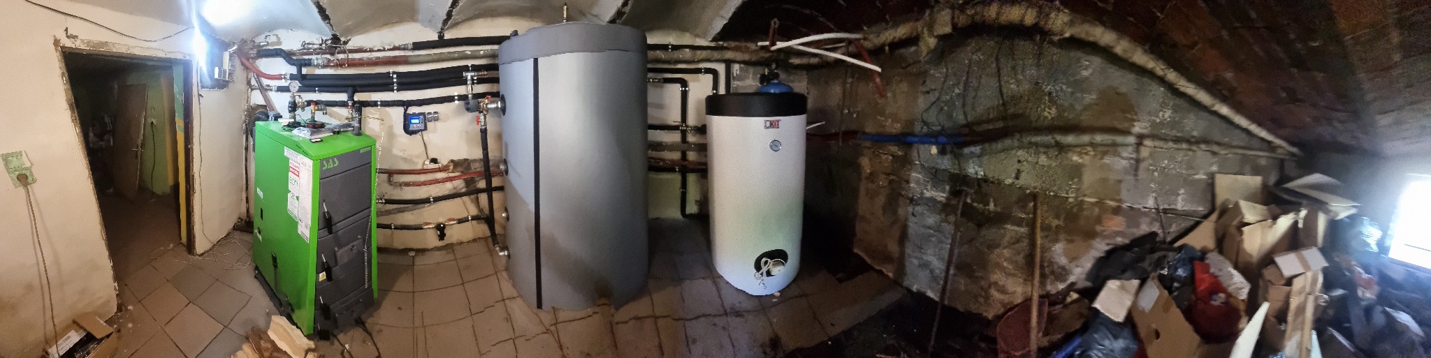 Widok panoramiczny kotłowni z zielonym kotłem, dwoma zbiornikami akumulacyjnymi, rurami i osprzętem, w pomieszczeniu z łukowym sufitem i częściowo odsłoniętą cegłą.