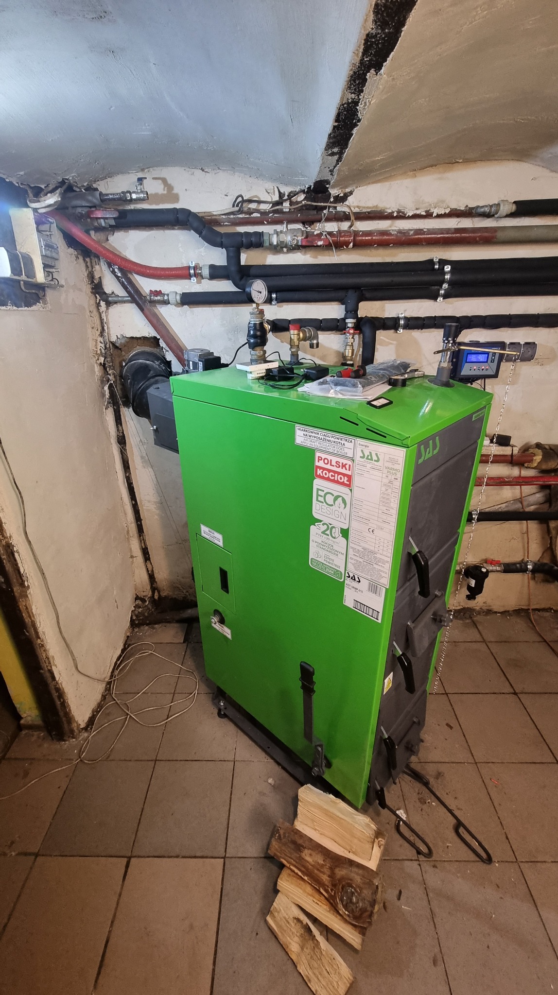Zielony kocioł na paliwo stałe marki SAS w piwnicy, z widocznymi rurami instalacji grzewczej i stosem drewna obok.