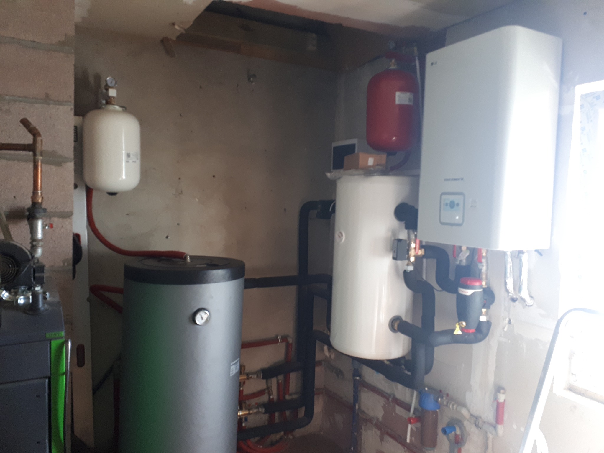 Instalacja grzewcza w kotłowni: szary zbiornik akumulacyjny, biały zasobnik ciepłej wody, biała pompa ciepła z widocznymi rurami i osprzętem, oraz czerwony zbiornik wyrównawczy.