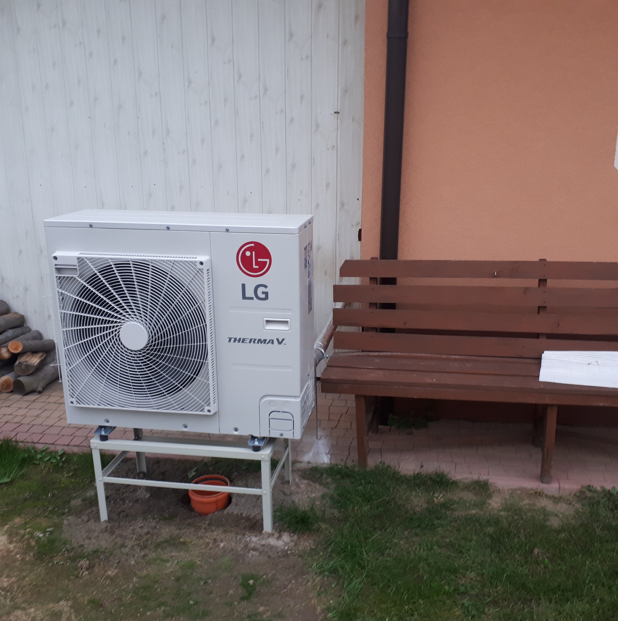 Zewnętrzna jednostka pompy ciepła LG Therma V na metalowym stelażu, obok drewniana ławka, na tle jasnej ściany z desek i fragmentu brązowej elewacji.