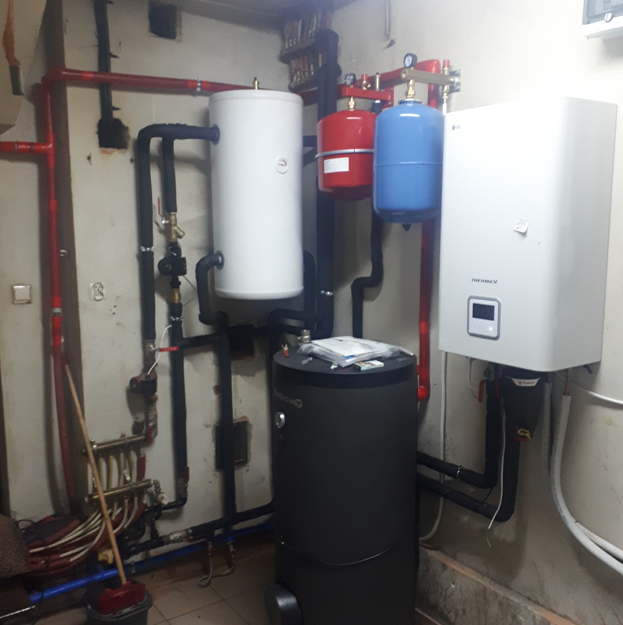 Zestaw urządzeń grzewczych w kotłowni: biały zbiornik buforowy, czerwony i niebieski zbiornik wyrównawczy, czarny zasobnik CWU i biała pompa ciepła LG Therma V, połączone rurami w systemie grzewczym.