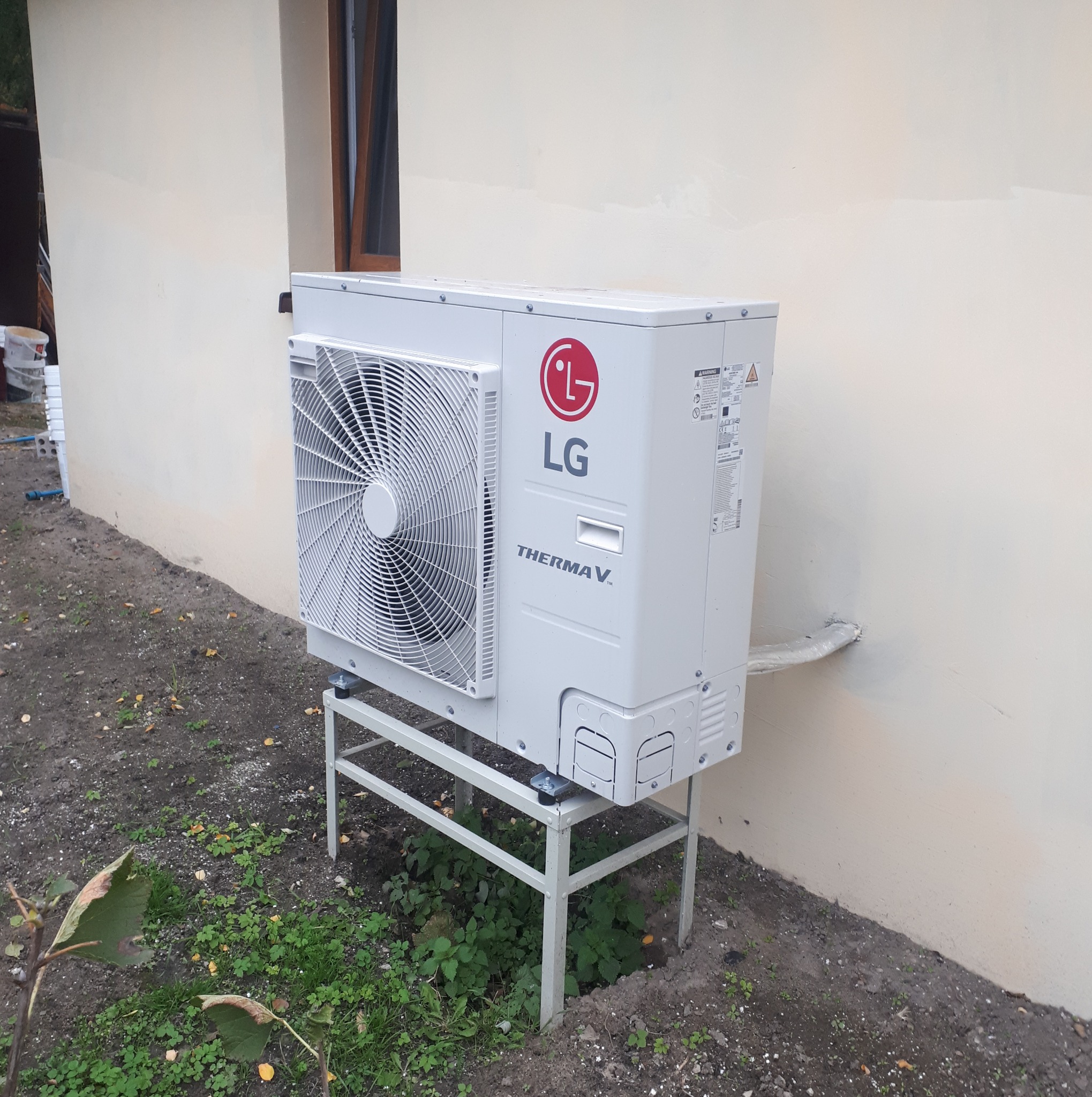 Biała jednostka zewnętrzna pompy ciepła LG Therma V, zamontowana na metalowym stelażu przy jasnej ścianie budynku z widocznym oknem.