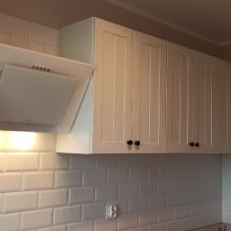 Wykończenia Przemysław Robert - Białe, stylizowane na postarzane, wiszące szafki kuchenne z czarnymi gałkami, podświetlona biała okap kuchenny i białe, prostokątne płytki na ścianie.