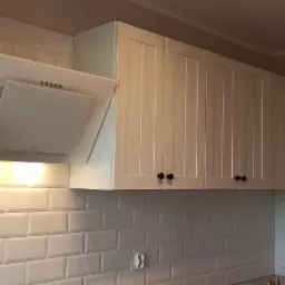 Białe, stylizowane na postarzane, wiszące szafki kuchenne z czarnymi gałkami, podświetlona biała okap kuchenny i białe, prostokątne płytki na ścianie.